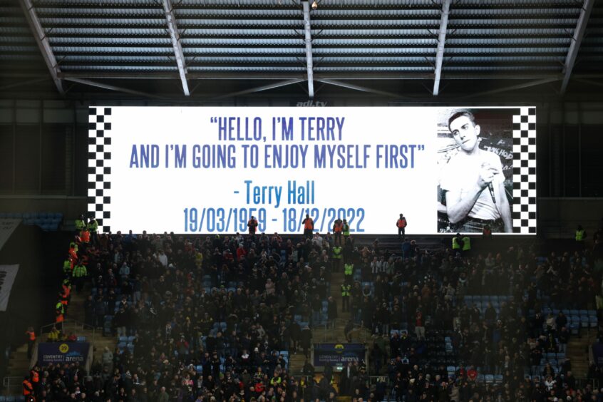 large screen at a footnall stadium featuring a photo of Terry Hall and the message 'Hello, I'm Terry and I'm going to enjoy myself first. Terry Hall 19/03/1959 - 18/12/2022.'