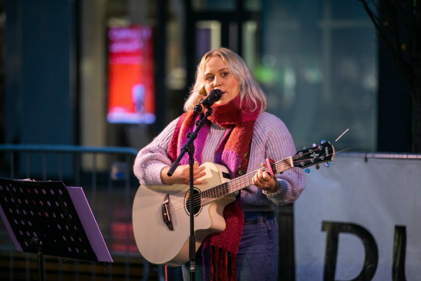 Katie Nicol plays and acoustic guitar on an outdoor stage, wearing a pink jumper and pink-and-orange scarf.