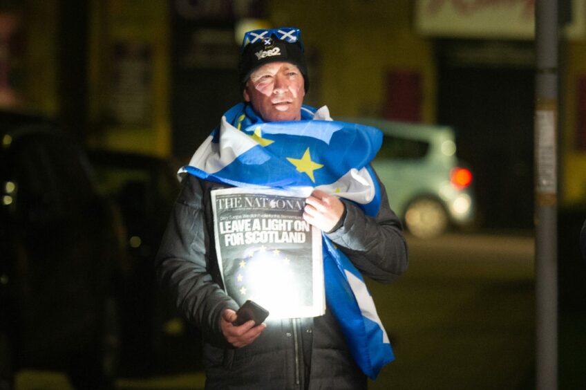 man draped in EU flag shining his mobile phone's torch on a front page which reads 'Leave a light on for Scotland'.
