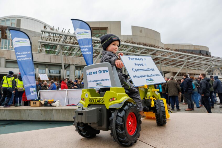 small boy on a toy tractor holding a banner which reads 'Farmers = food = communities' and a crowd of protesters outside the Scottish parliament.
