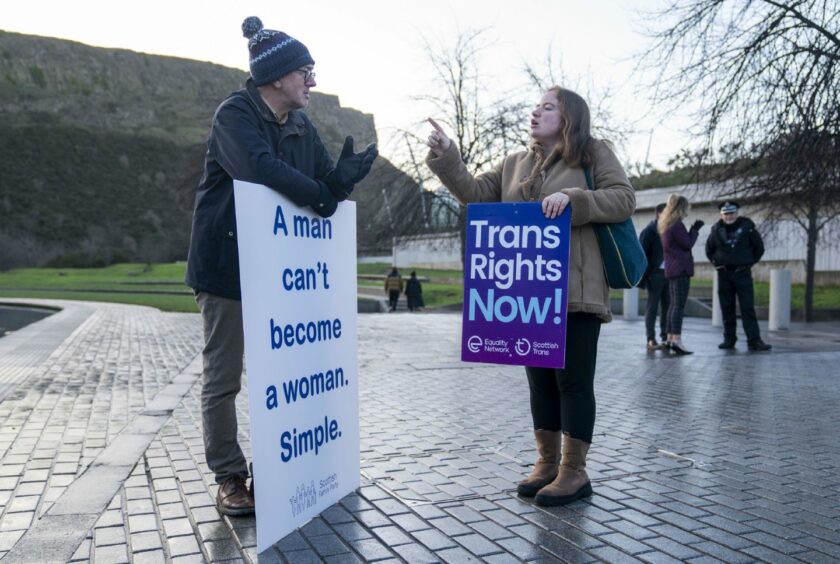 Man and woman arguing outside Scottish parliament. Man holds a placard which reads 'A man can't become a woman. Simple'. Woman holds a placard which reads 'Trans rights now'.