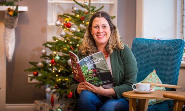 A smiling woman holding The Perthshire Cookbook sitting in front of a Christmas tree