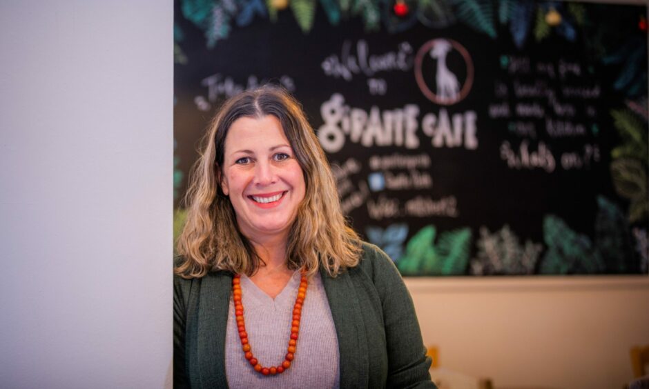 A smiling woman in a green cardigan, grey top and orange necklace in front of a sign that says welcome to the Giraffe Cafe