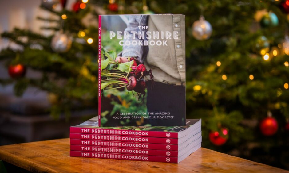 A stack of The Perthshire Cookbook, with a farmer holding radishes on the cover, in front of a Christmas tree