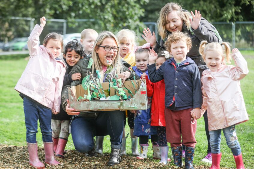 a group of nursery aged children in wellies waving happily next to a model forest