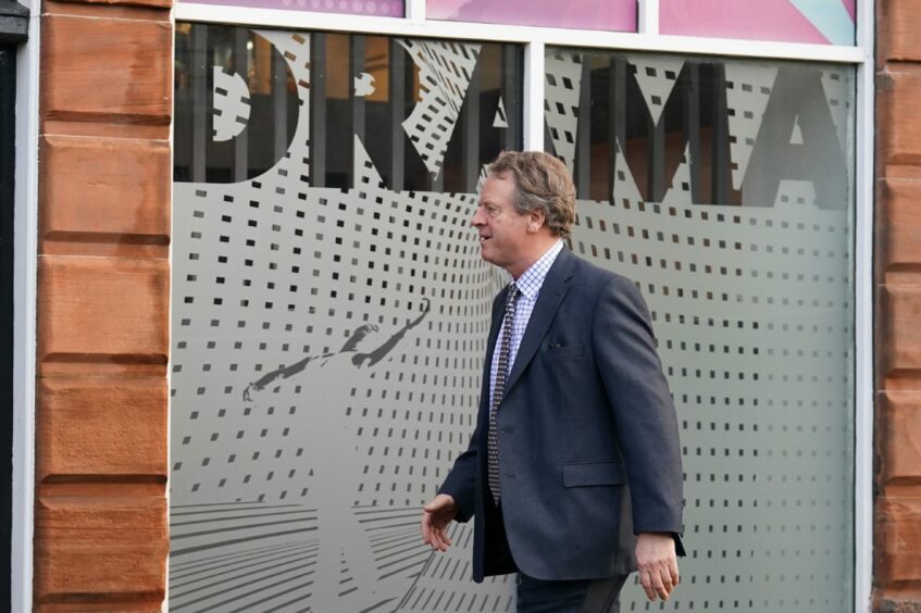 Scottish Secretary Alister Jack walking in front of a window with the word DRAMA spelled out in large letters.