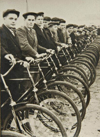 vintage photo of men in cloth caps lined up on bicycles.
