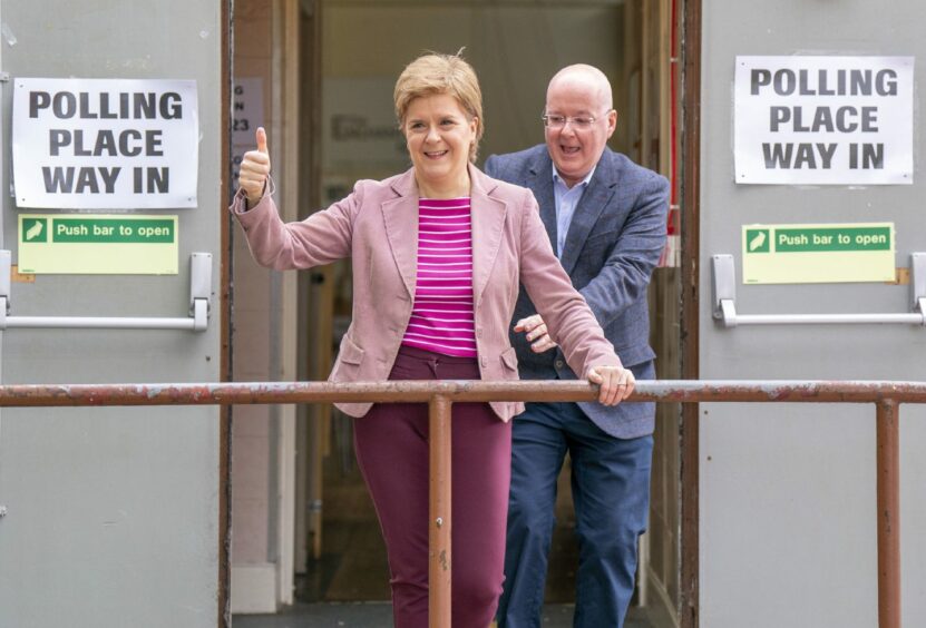 Nicola Sturgeon giving a thumbs up outside a polling station while her husband Peter Murrell emerges behind her.
