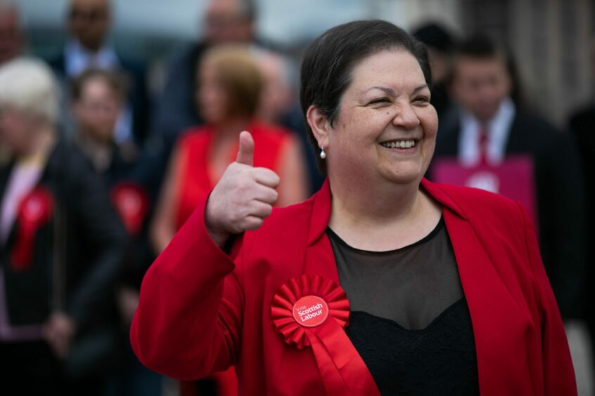 Jackie Baillie in red jacket with red Labour rosette giving a thumbs-up sign