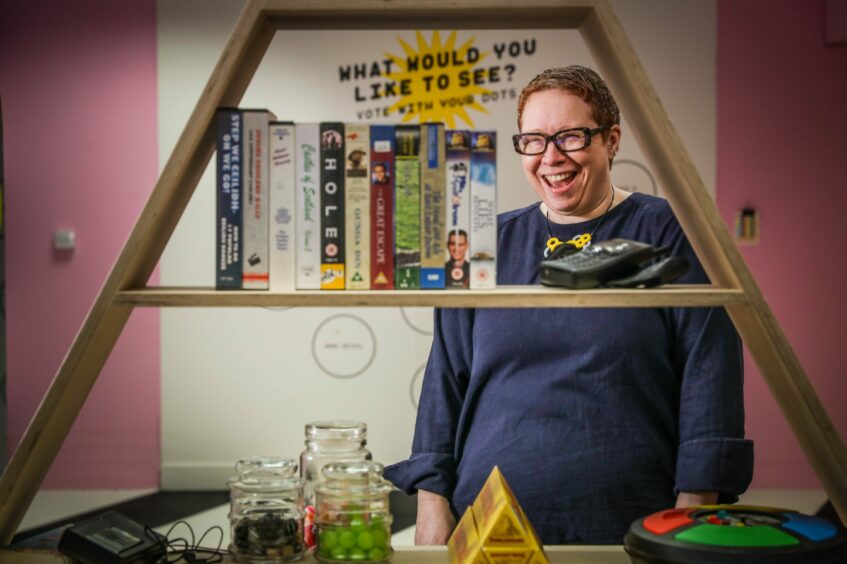 Donna Holford-Lovell next to a shelf, surrounded by objects in front of a wall with the words 'what would you like to see?' written on it