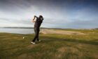 Golfer taking a swing with Scottish beach in background.