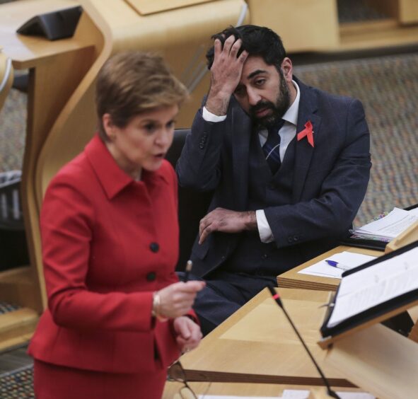 Nicola Sturgeon speaking in the Scottish parliament, watched by Humza Yousaf who is scratching his head.