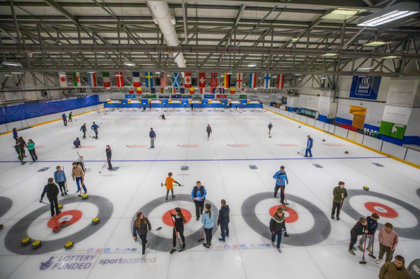 Overview of Dewars Centre interior, Perth, with national flags draped in background and young curlers practising on the ice