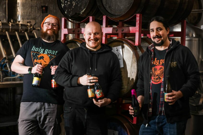 Three men stand in front of barrels of beer holding bottles of Holy Goat beer.