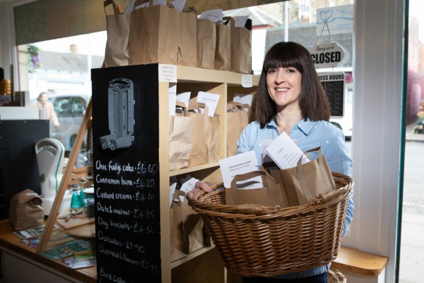 A dark-haired woman stands in a storefront, with a basket full of brown paper bags, which contain baking ingredients. 