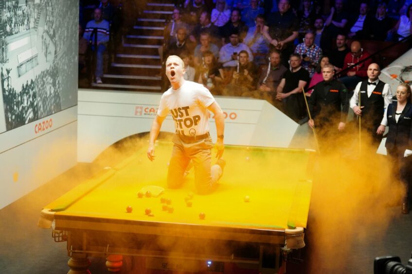 A man in a Just Stop Oil T shirt kneeling on a snooker table in a cloud of orange power, during a match at the World Snooker Championship at the Crucible Theatre, Sheffield,