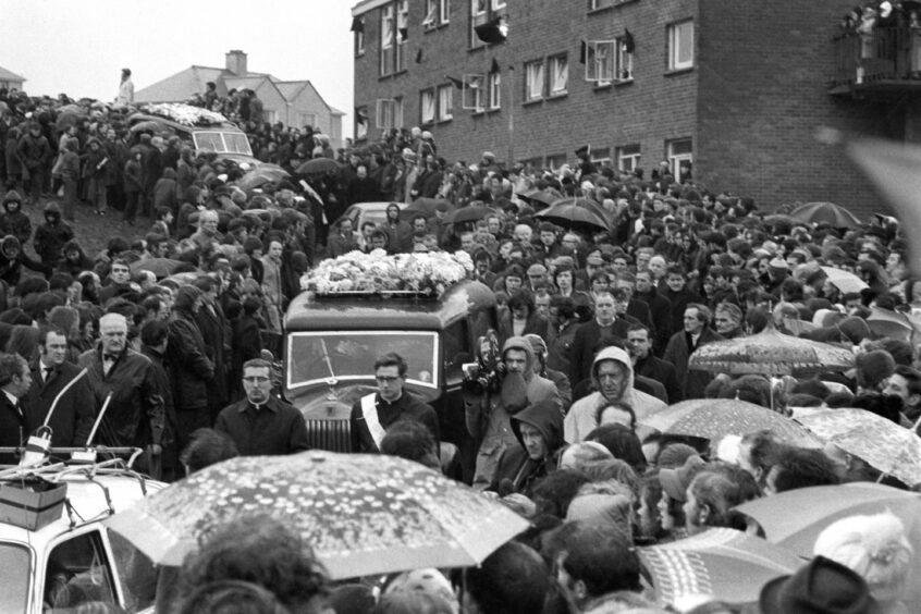 A silent crowd form a cordon 10 or more deep, lining both sides of the 250 yards of road leading from St Mary's Church, Creggan Hill to the cemetery, to watch the funeral procession of the 13 who died on "Bloody Sunday" in 1972.