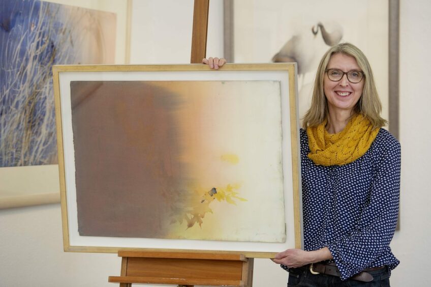 The artist Claire Harkess is standing beside one of her paintings. She is wearing a blue jumper and yellow scarf and is smiling.