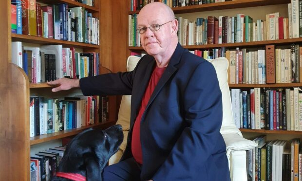 Author David O'Donnell is pictured sitting on a white armchair with a black Labrador dog. David is wearing a navy suit and red top and there are well-stocked bookshelves in the background.