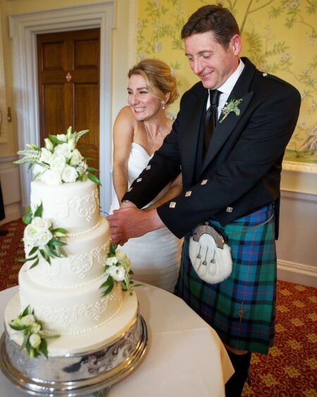 Laura, in a wedding dress, and Steven, in a kilt, cutting their three tier wedding cake.