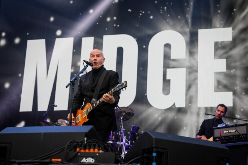 Midge Ure playing guitar on stage in front of a backdrop which reads 'MIDGE' in large letters.