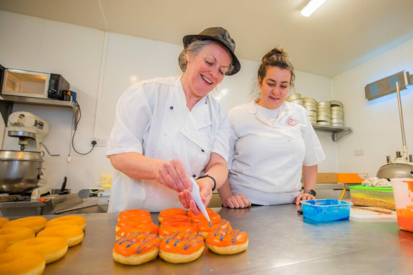 Two female bakers in the kitchen, decorating orange doughnuts with a blue glaze.