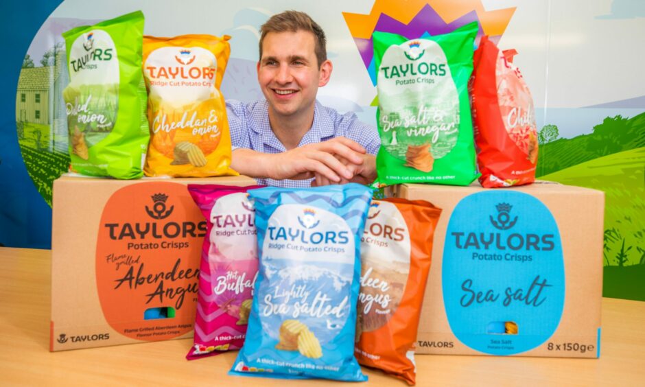A man sitting down surrounded by bags and boxes of Taylors Snacks crisps.