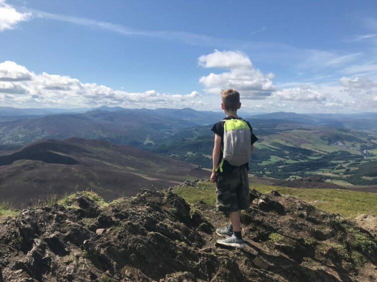 A young boy stands on the top of a mountain with a blue sky beyond on a day of walking in Highland Perthshire. 