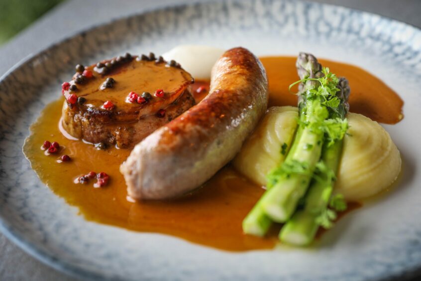 A grey plate with pork belly, sausage, clotted cream mash, asparagus and peppercorn sauce