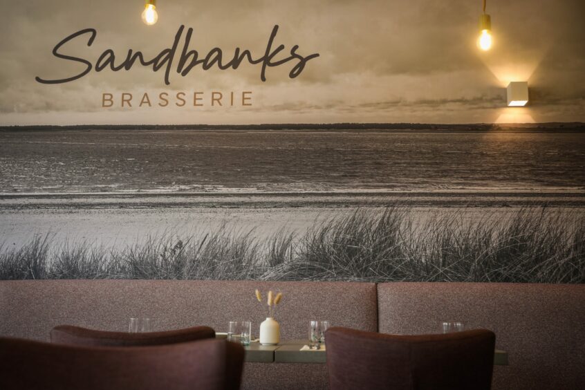 interior of Sandbanks brasserie in Broughty Ferry, showing table and chair with large mural depicting Broughty Ferry beach, Tay estuary and Tentsmuir beach and forest on far side.