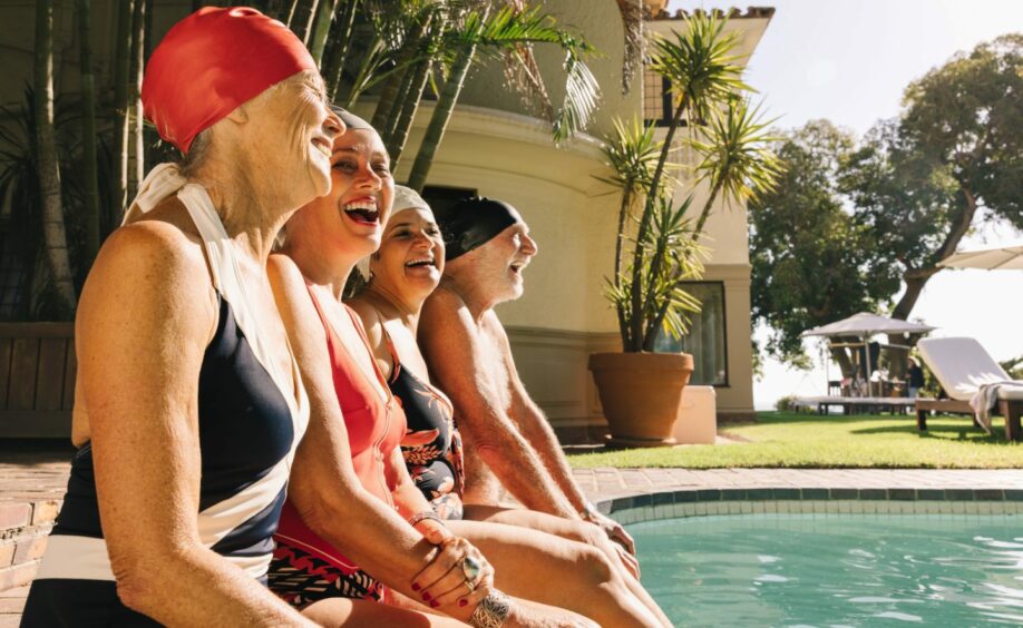 Four elderly people in swim suits laughing by a poolside.