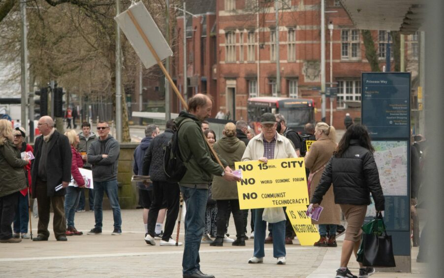 Man in city centre holding placard saying 'No 15-minute city, no World Economic Forum, no Communist technocracy'.