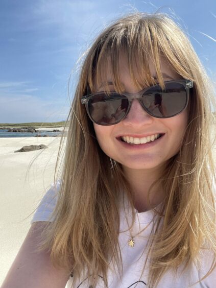 Rebecca Baird smiling broadly in large sunglasses on North Beach Iona, against white sands and a bright blue sky.