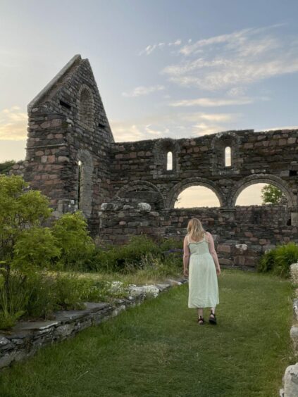 Rebecca Baird in green maxi dress from behind, exploring ruins of the old nunnery on Iona as the sun sets behind her.