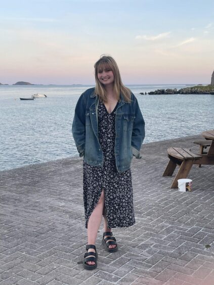 Rebecca Baird in floral maxi dress and denim jacket smiling as she poses in a pub beer garden next to the sea at sunset.