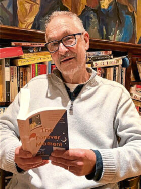 The image shows author Paul A Mendelson reading from a copy of The Forever Moment.