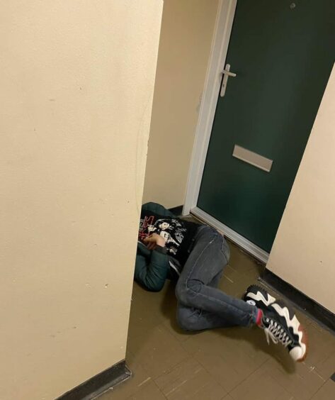 A man with his face hidden behind a wall, lying on the landing of a block of flats