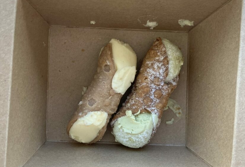 Two cannolis, one with yellow and one with green filling, inside a brown takeaway box.