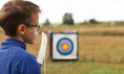 Child camper at Over the Wall camp taking part in archery. He has a breathing tube attached to his face. He looks happy and engaged.