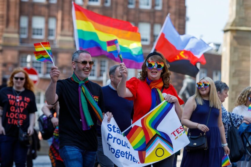 People marching through Dundee as part of the weekend's Pride celebrations. A man and a woman in the foreground are waving rainbow flags and carrying a banner which reads 'Standing proud together'.