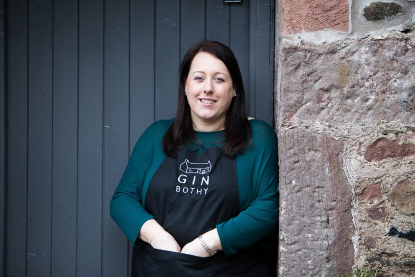 A woman standing outside a bothy wearing a black apron saying Gin Bothy.