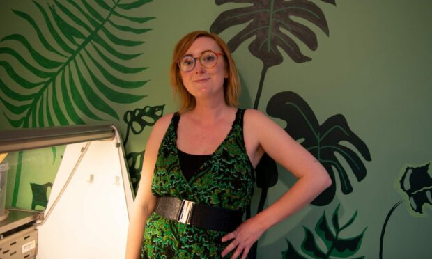 A woman wearing rainbow glasses and a green jumpsuit in front of the takeaway counter in The Little Green Larder.
