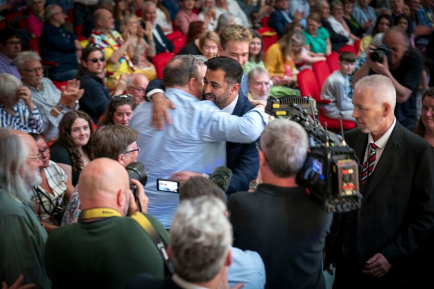 First Minister Humza Yousaf embraces a man in the audience at the SNP independence convention at the Caird Hall in Dundee. The pair are being filmed and photographed by members of the media as audience members applaud.