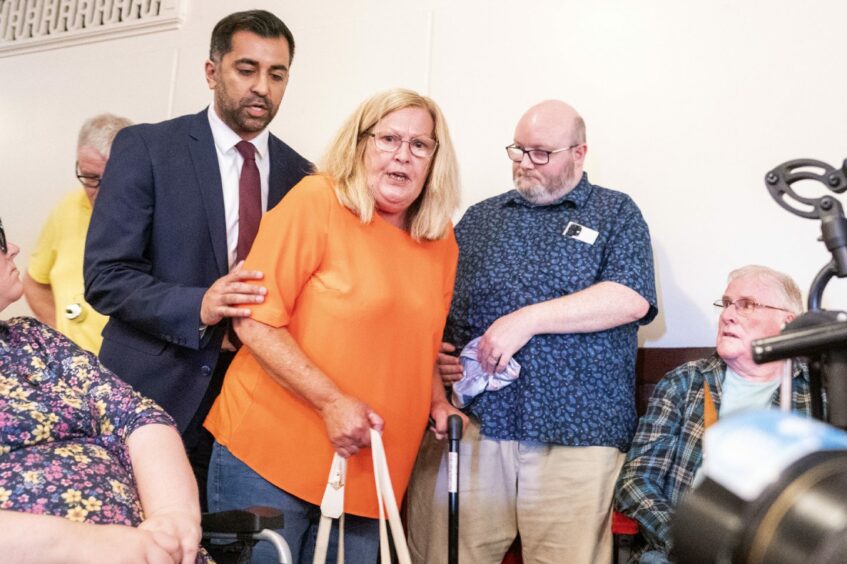 First Minister Humza Yousaf with his hand on the arm of Theresa Mallet, who is walking with a stick. Ms Mallet is talking as the pair walk past a number of people in the Caird Hall, Dundee.