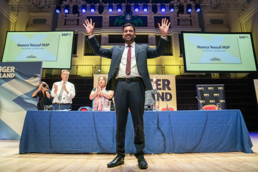 Humza Yousaf holding up both hands to the audience on stage at the Caird Hall in Dundee. A number of supporters stand applauding at a table on the stage behind him. 