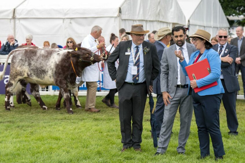 A suited First Minister Humza Yousaf talking to officials in the ring at the Highland Show while an exhibitor in white coat walks a cow behind them.