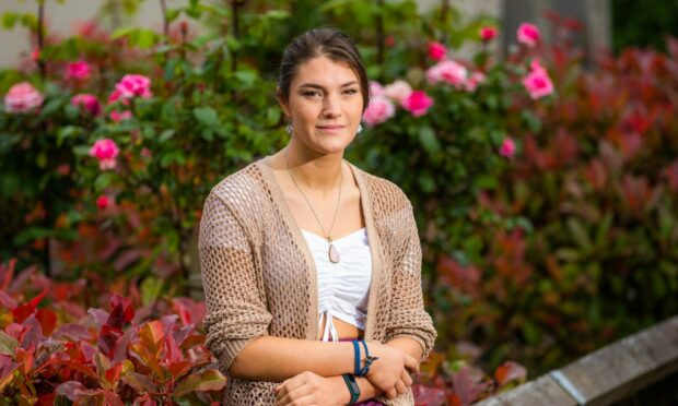 Picture shows Briony Paddon wearing a white top and brown cardigan sitting in front of a bush of pink roses.