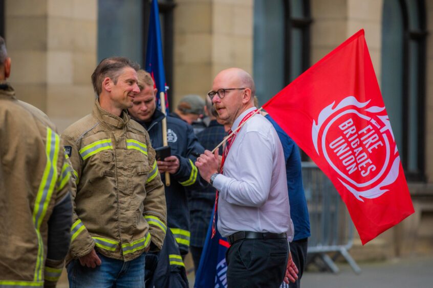 David Evans in firefighter uniform talking to man in shirt and tie holding a Fire Brigades Union flag.