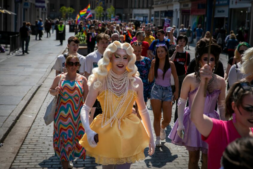Large crowd of people walking through Dundee city centre for Pride. A smiling drag queen in yellow satin dress and long blonde hair wearing many strands of pearls is front and centre.