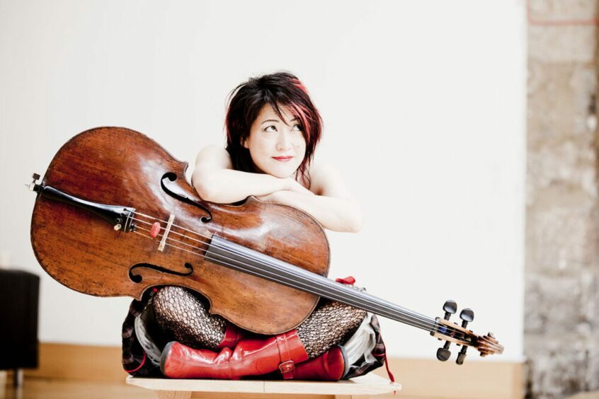 Cellist Su-a Lee is pictured sitting cross-legged on the floor holding her cello.